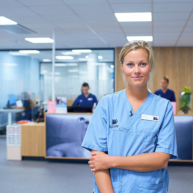 Kvinnlig veterinär iklädd ljusblåa vårdkläder ler mot kameran i en djurklinik. Bakom henne syns en reception där två personer står och jobbar.