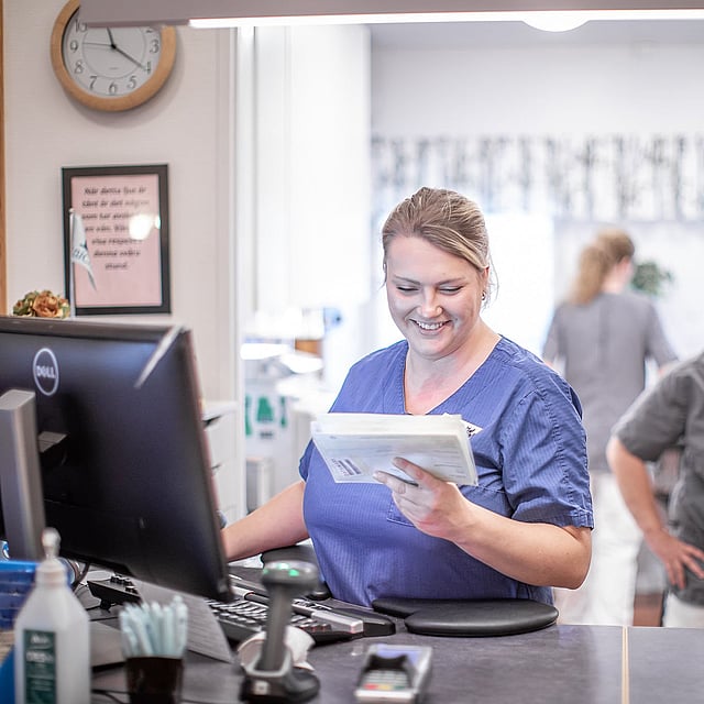 Kvinnlig veterinär står framför en dator och kollar ner mot en anteckningsblock och ler. Bakom henne syns en till glad kvinnlig veterinär och bakom dem finns det ännu en veterinär som står med ryggen mot alla.