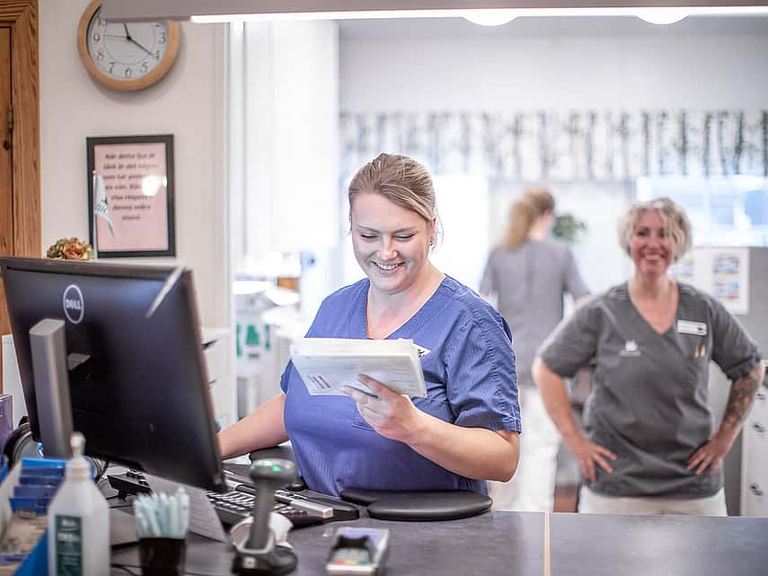 Kvinnlig veterinär står framför en dator och kollar ner mot en anteckningsblock och ler. Bakom henne syns en till glad kvinnlig veterinär och bakom dem finns det ännu en veterinär som står med ryggen mot alla.