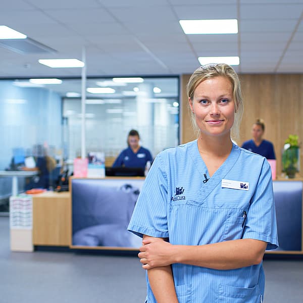 Kvinnlig veterinär iklädd ljusblåa vårdkläder ler mot kameran i en djurklinik. Bakom henne syns en reception där två personer står och jobbar.