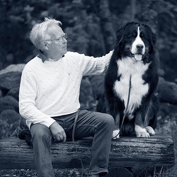 En äldre man sitter på en stock med sin stora Berner sennenhund ute i naturen. Mannen klappar på hunden. Bilden är i svartvitt.