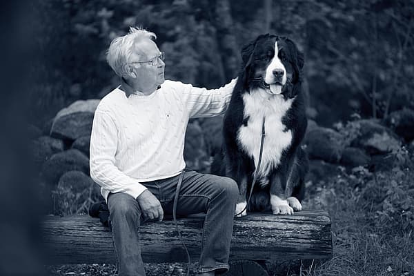 En äldre man sitter på en stock med sin stora Berner sennenhund ute i naturen. Mannen klappar på hunden. Bilden är i svartvitt.